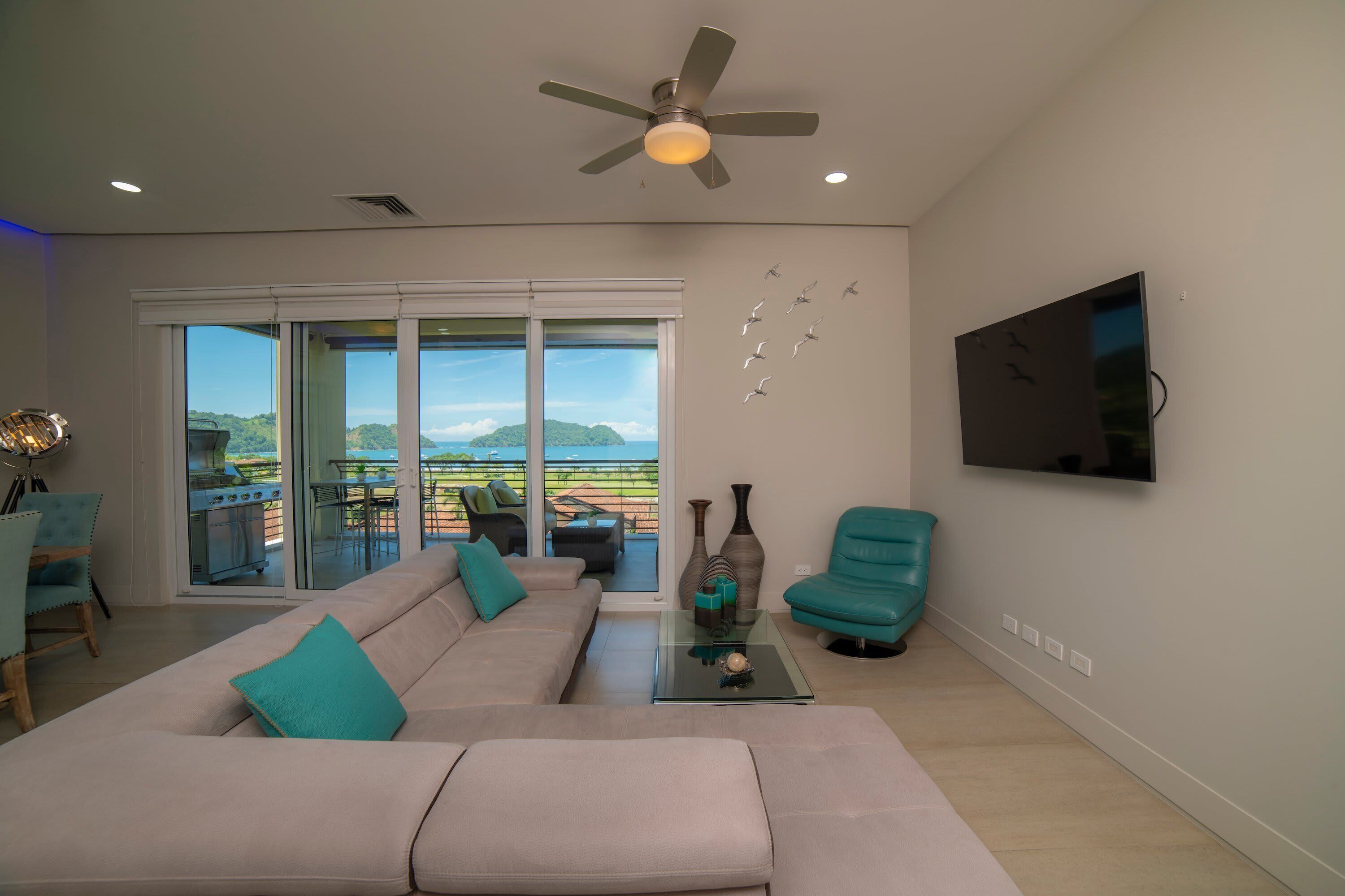 Ein Wohnzimmer mit Sofa, Sesseln, einem Couchtisch, Deckenventilator, Fernseher und Blick durch Glastüren.