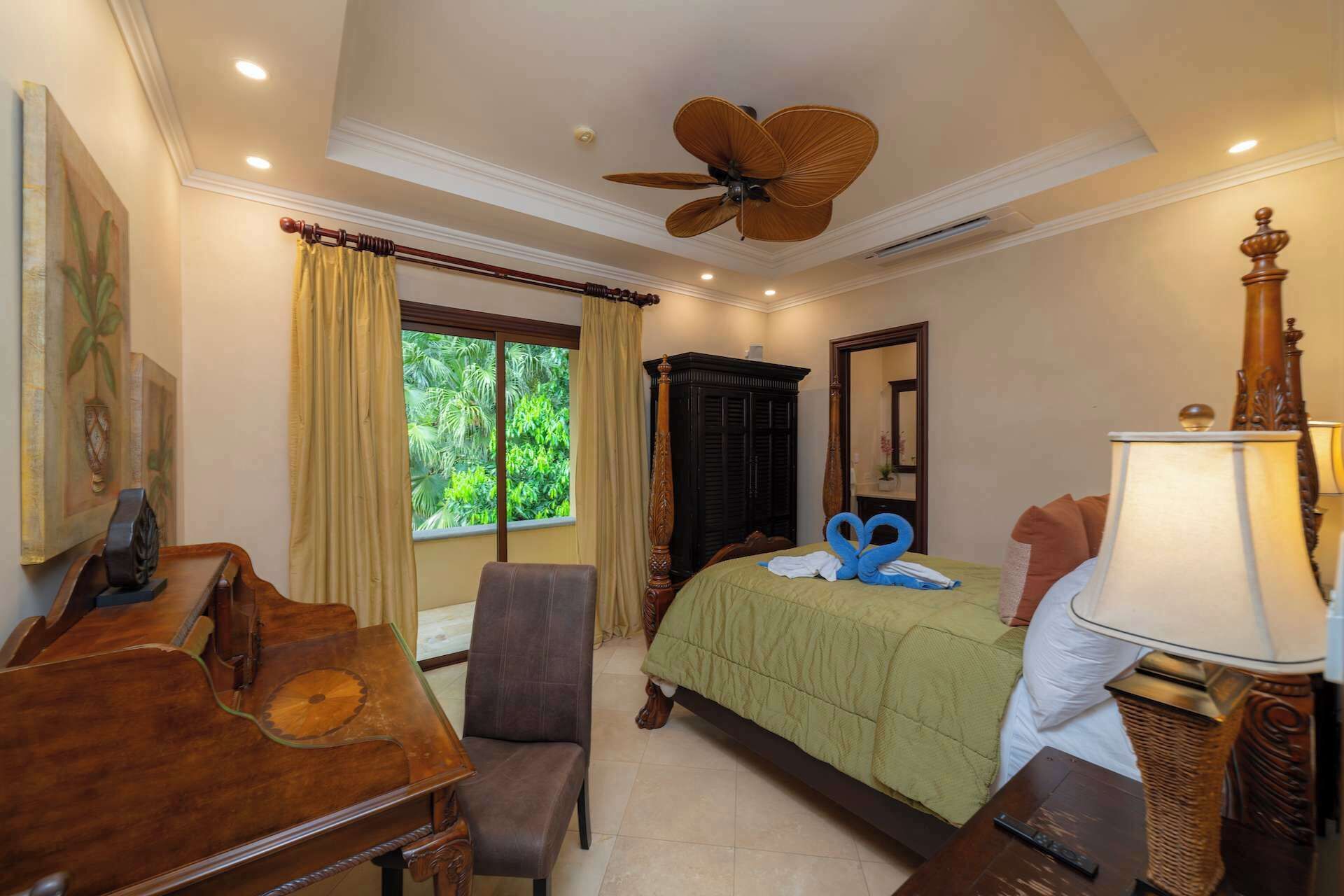 A hotel room with a bed, a desk, a chair, a ceiling fan, a window with curtains, and a lamp.