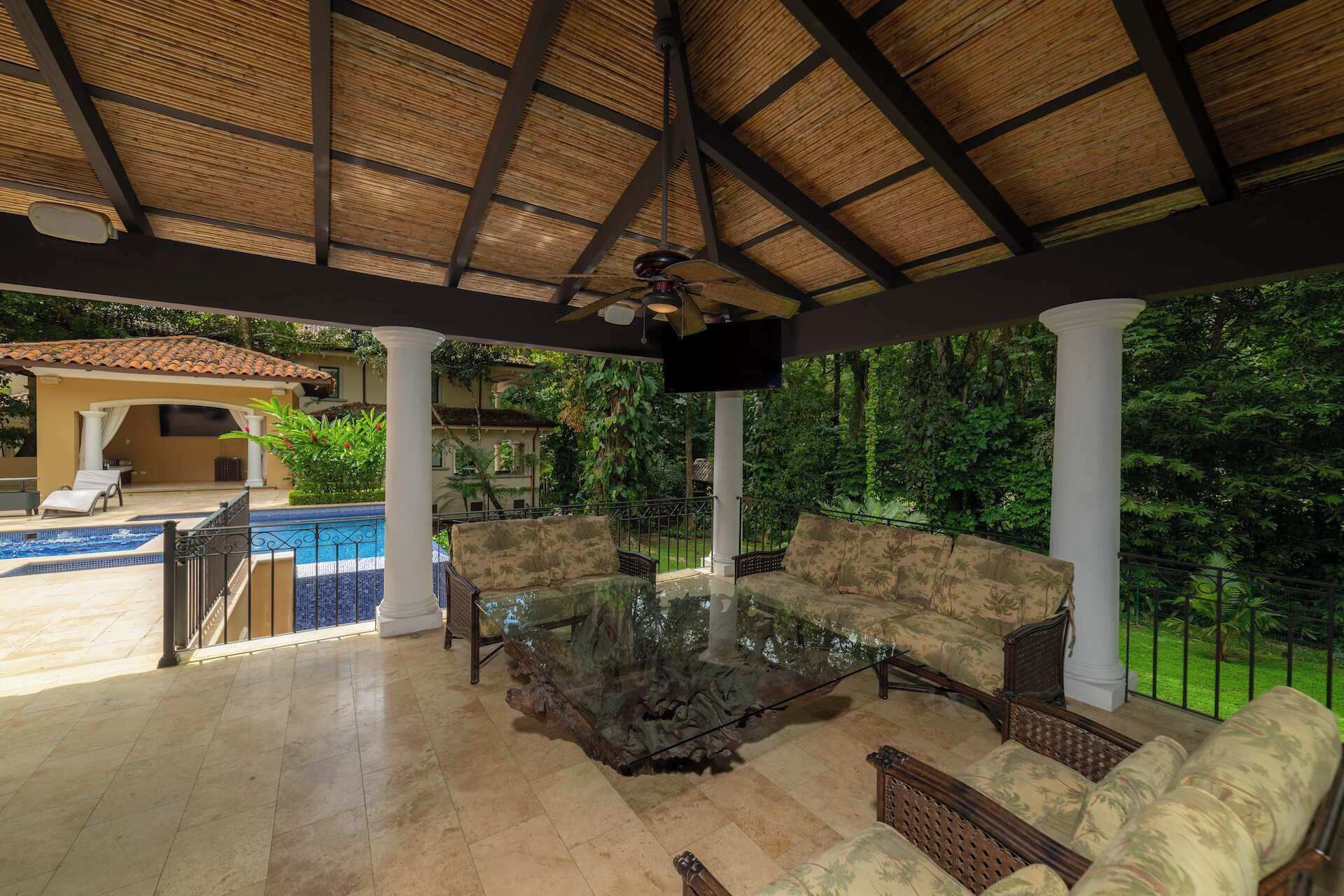 A covered outdoor seating area with a pool, a TV, and lush greenery.
