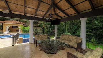 A covered outdoor seating area with a pool, a TV, and lush greenery.