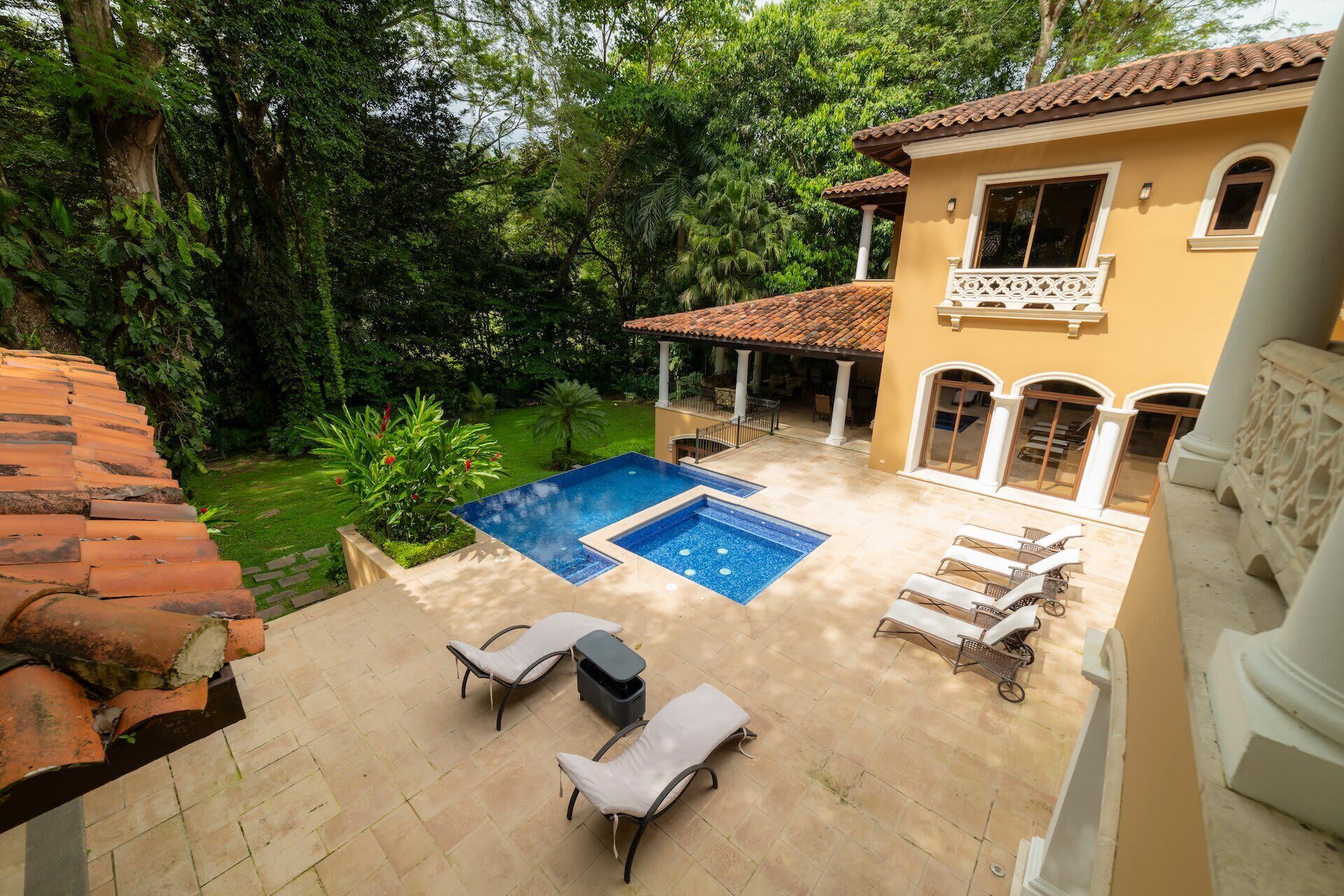 Une piscine avec des chaises longues, un patio couvert et un jardin arboré.