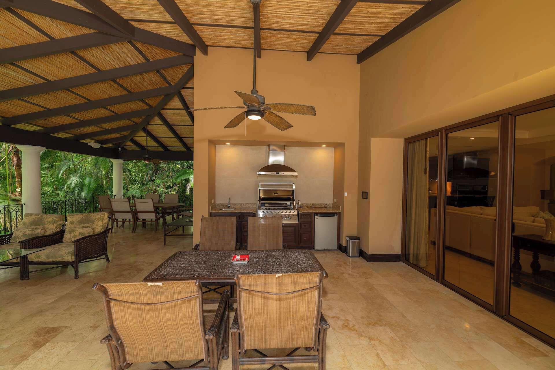 An outdoor kitchen area with a dining table and chairs, a ceiling fan, and a view of the surrounding greenery.
