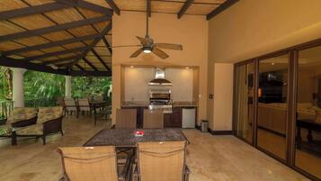 An outdoor kitchen area with a dining table and chairs, a ceiling fan, and a view of the surrounding greenery.