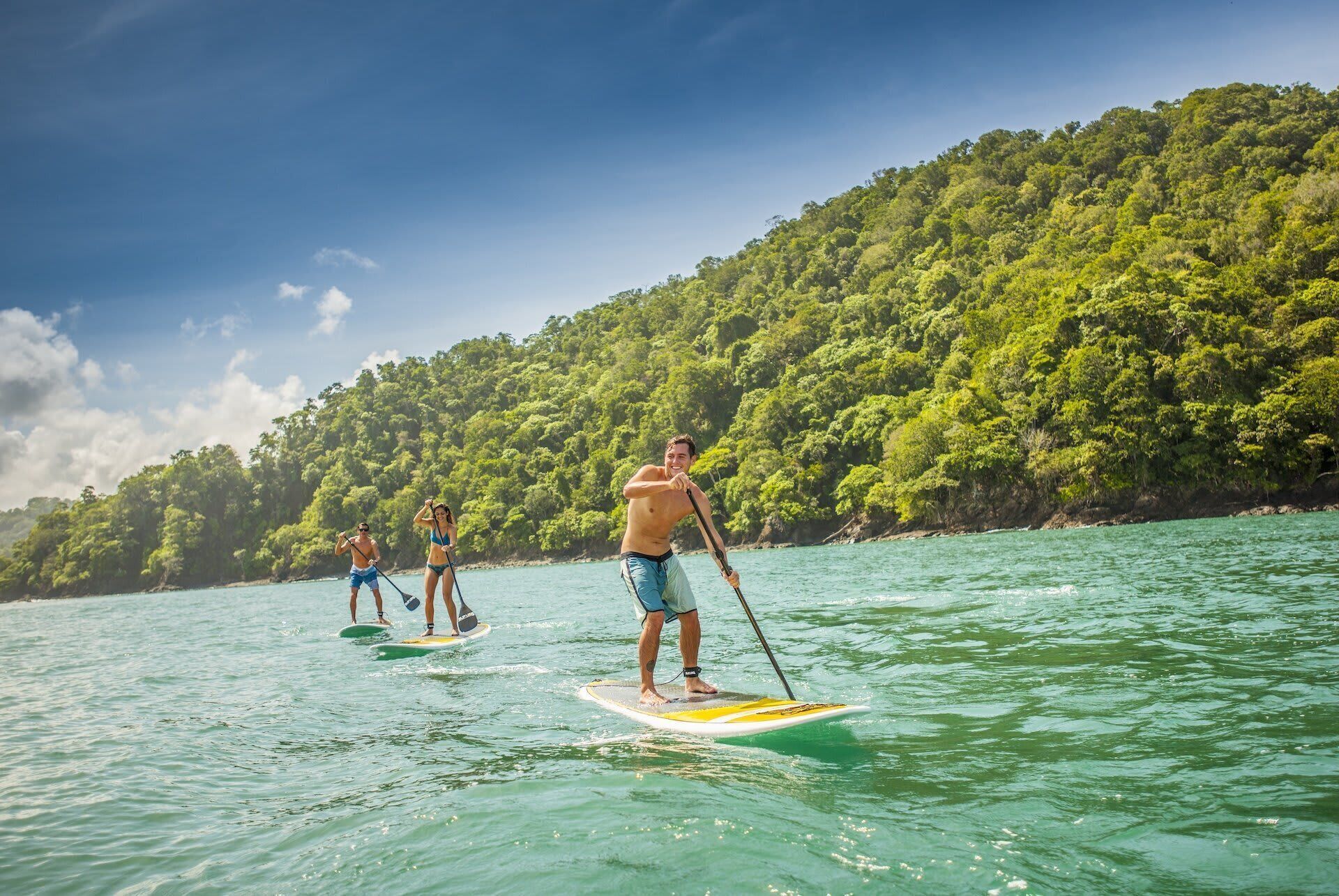 Tres personas haciendo paddleboard en un mar tranquilo, con una colina boscosa al fondo.