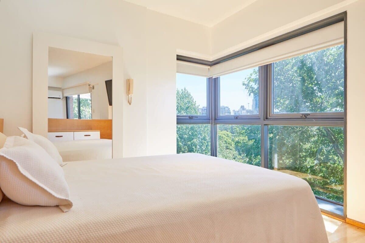 A bedroom with a large window, a bed, a bedside table, and a mirror.