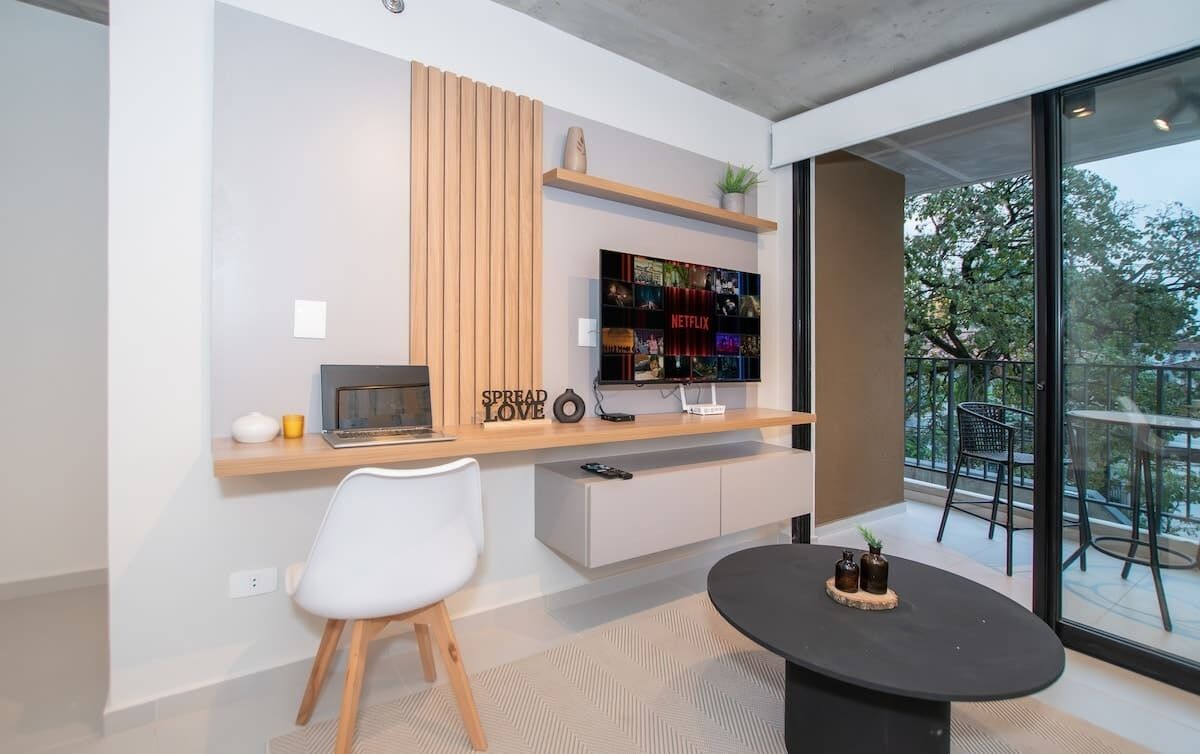 Ein modernes Wohnzimmer mit Fernseher, einer Holzreolenwand, einem weißen Sessel, einem schwarzen Tisch und einem Balkon mit Gartenmöbeln.