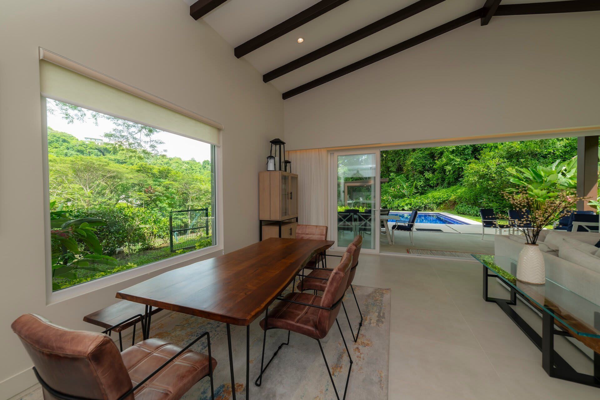 A spacious room with a large dining table, comfortable seating, a glass door leading to a pool area, and a view of lush greenery.