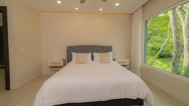Een slaapkamer met een bed, nachtkastjes, een plafondventilator, inbouwverlichting en een raam met uitzicht op bomen.