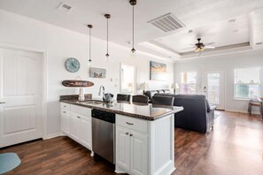Eine geräumige Küche mit weißen Schränken, einer Granit Arbeitsplatte und einem Geschirrspüler. Es gibt einen Essbereich mit einer dunkelgrauen Couch und einem Deckenventilator.