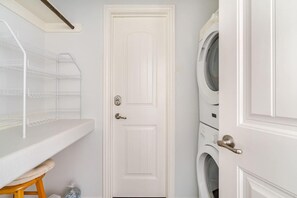A laundry room with a white door, a washing machine, and a shelf with hanging clothes. - Sarenity - 3Bd 2Bth, 8 Guests, Pet Friendly (Bolivar Peninsula)