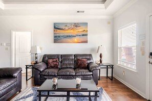 Un salon avec un canapé en cuir noir, une table basse en verre et un tableau représentant un coucher de soleil sur l’océan.
