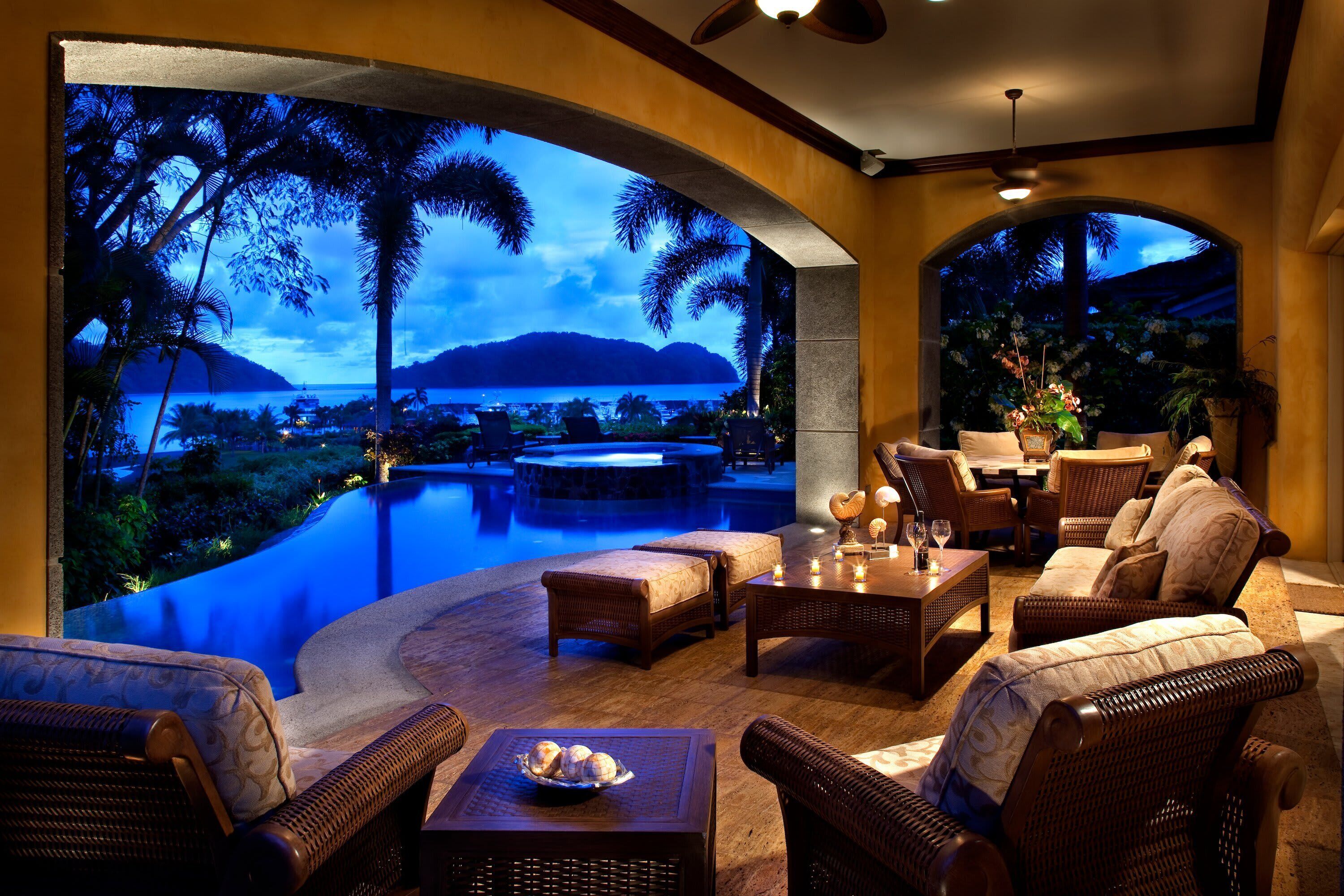 A poolside area with wicker furniture, a dining table, and a view of a tropical landscape with a pool and palm trees.