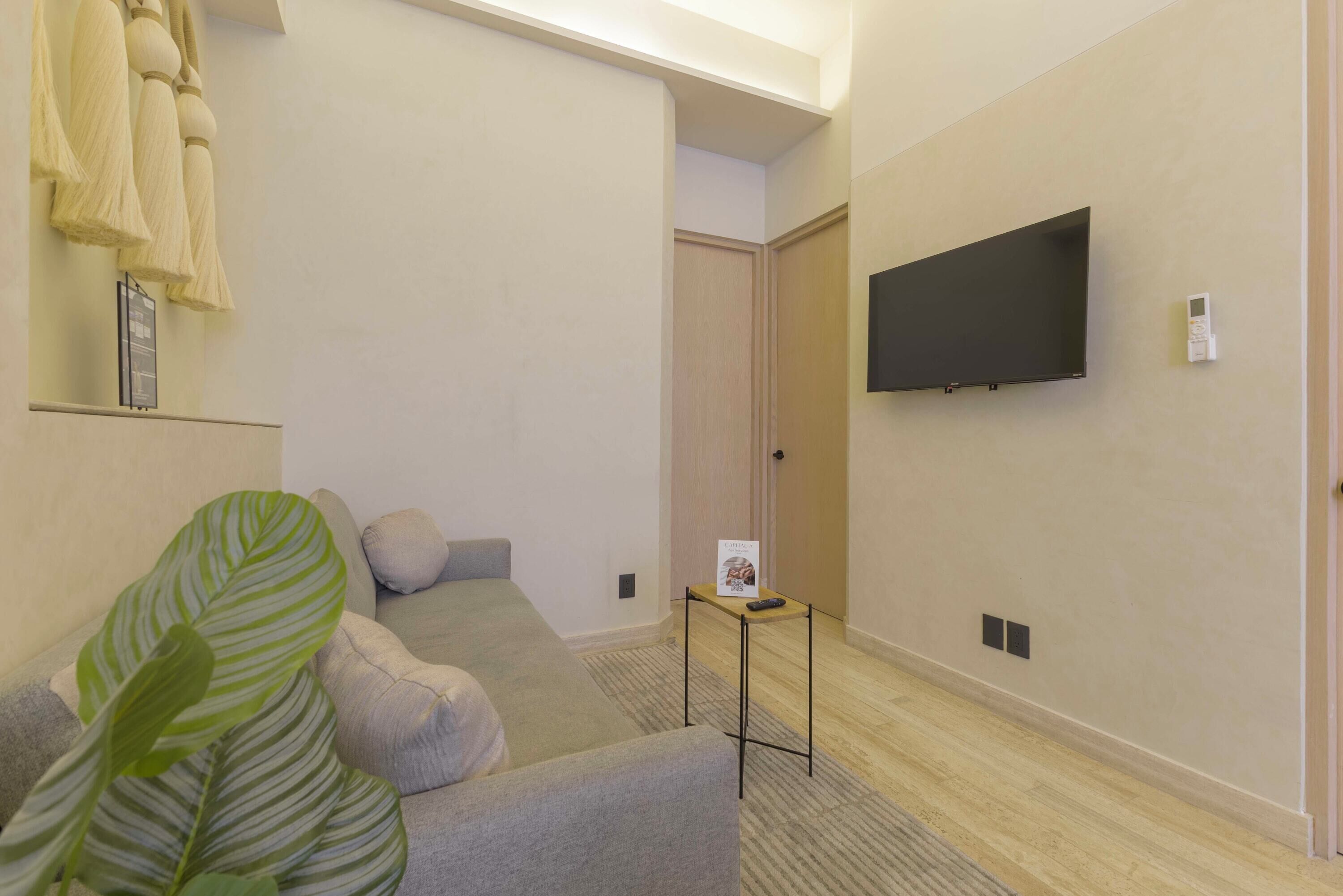 A hotel room with a sofa, a small table, a TV mounted on the wall, and a plant.
