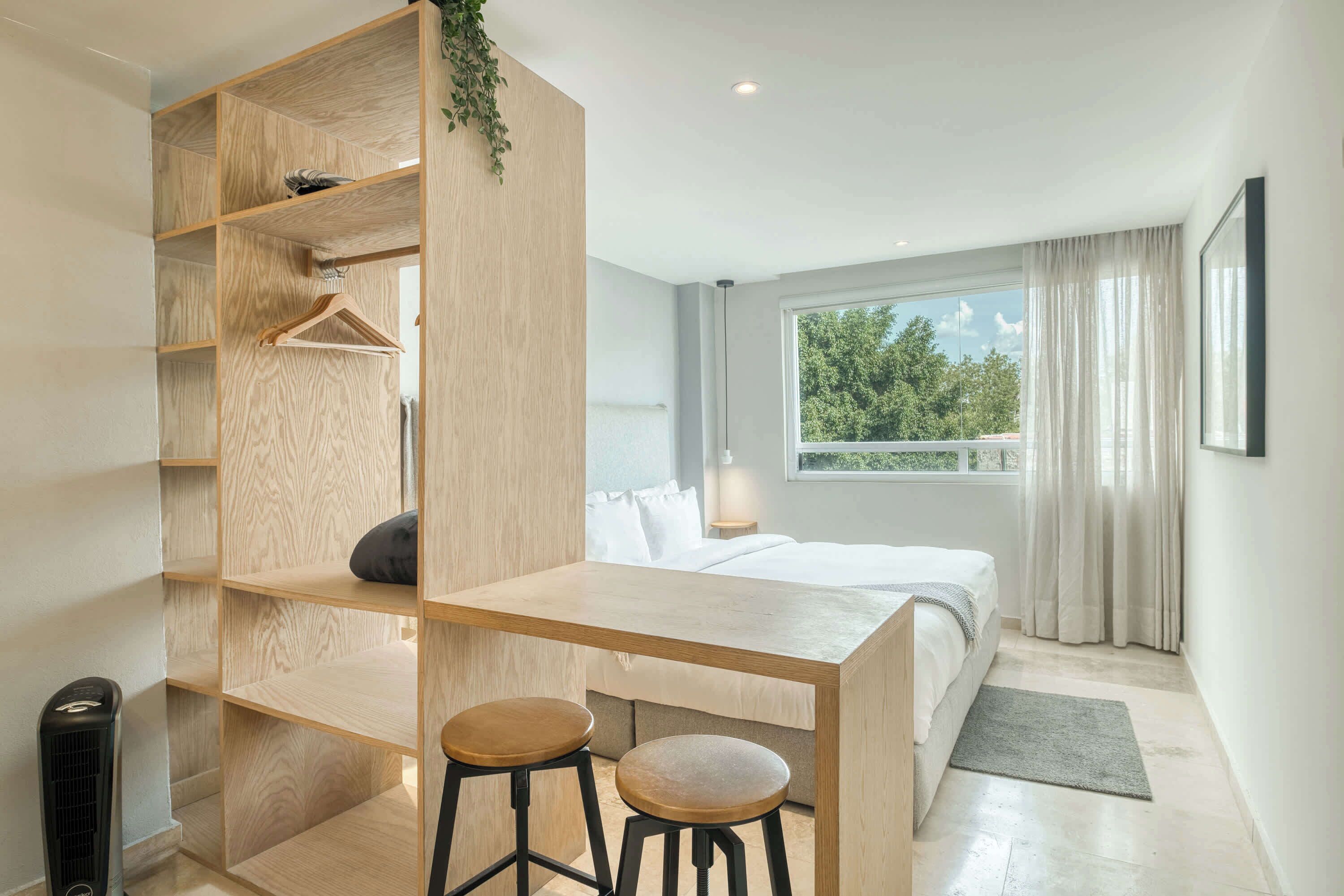 Une chambre à coucher comprenant un lit, un bureau avec deux tabourets, une armoire et une fenêtre donnant sur un paysage verdoyant.