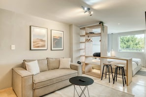 Ein modernes Wohnzimmer mit einer Couch, einem Couchtisch, einem Schreibtisch mit zwei Hocker und einem Bett. An der Wand hängen zwei gerahmte Bilder, auf dem Schreibtisch steht eine Pflanze und ein großes Fenster lässt natürliches Licht herein.