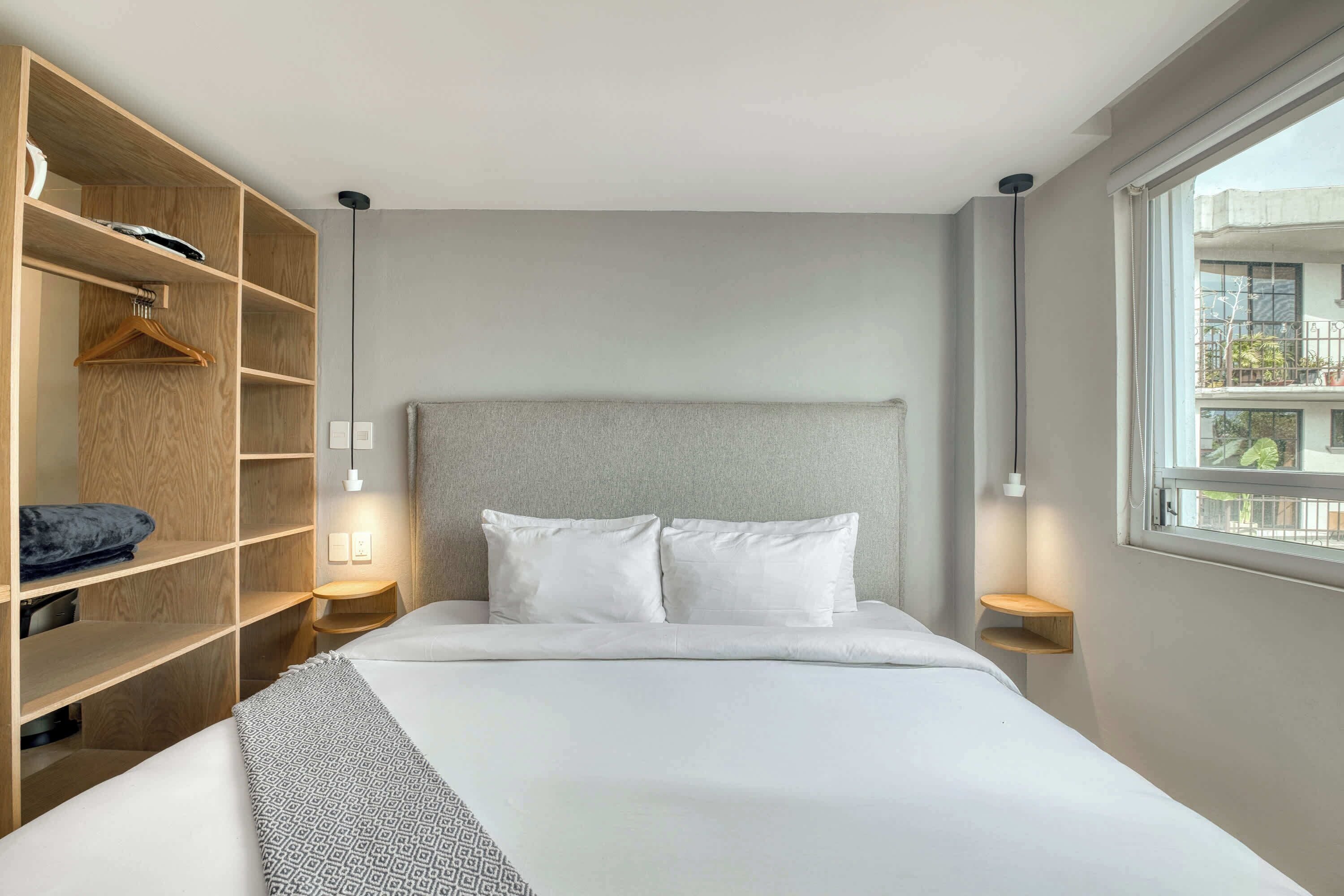 A modern hotel room with a large bed, a wooden wardrobe, bedside tables, and a window with a view.