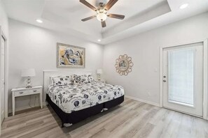 Une chambre à coucher comprenant un lit, des tables de chevet, un ventilateur de plafond, un tableau, un élément décoratif mural et une porte.