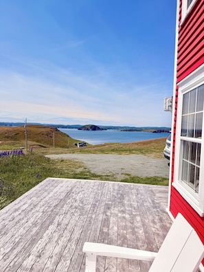 A wooden deck with a white chair, a red building, and a view of a lake and distant hills.