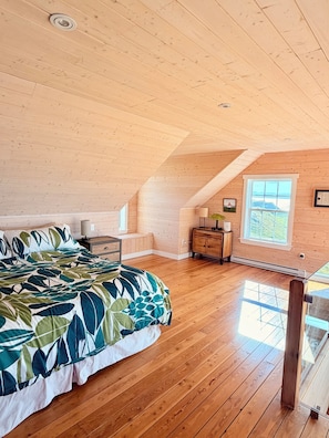 A bedroom with a large bed, wooden flooring, a dresser, a window with a view, and a sloped ceiling.