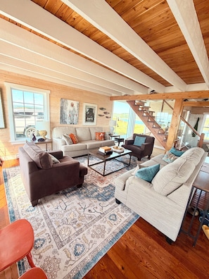 A spacious living room with a high ceiling, wooden beams, a large window, and a staircase leading to an upper level.
