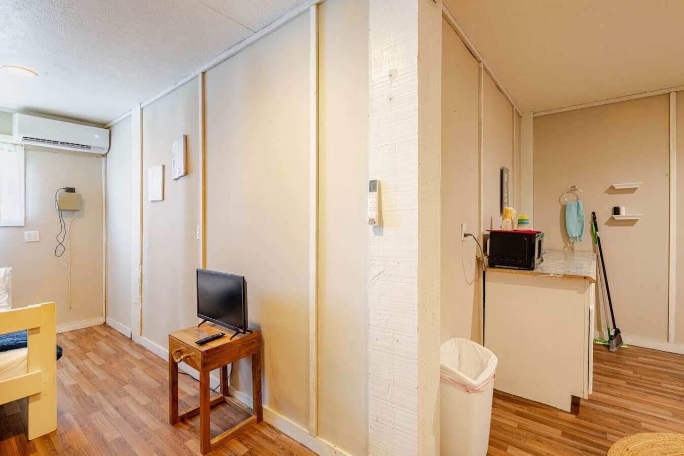 A room with a wooden floor, a small wooden table with a TV, a white trash bin, and a wall-mounted air conditioner.
