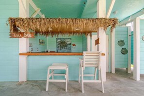 A bar area with a thatched roof, two white chairs, and a painting of a palm tree. - 3 King Suites,  15 Guests - Sunzout (Bolivar Peninsula)