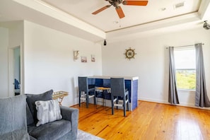 A living room with a grey sofa, wooden flooring, a ceiling fan, a small table with chairs, and a window with curtains. - 3 King Suites,  15 Guests - Sunzout (Bolivar Peninsula)