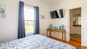Ein Schlafzimmer mit Bett, Fernseher, Fenster mit Vorhängen und Blick nach draußen.