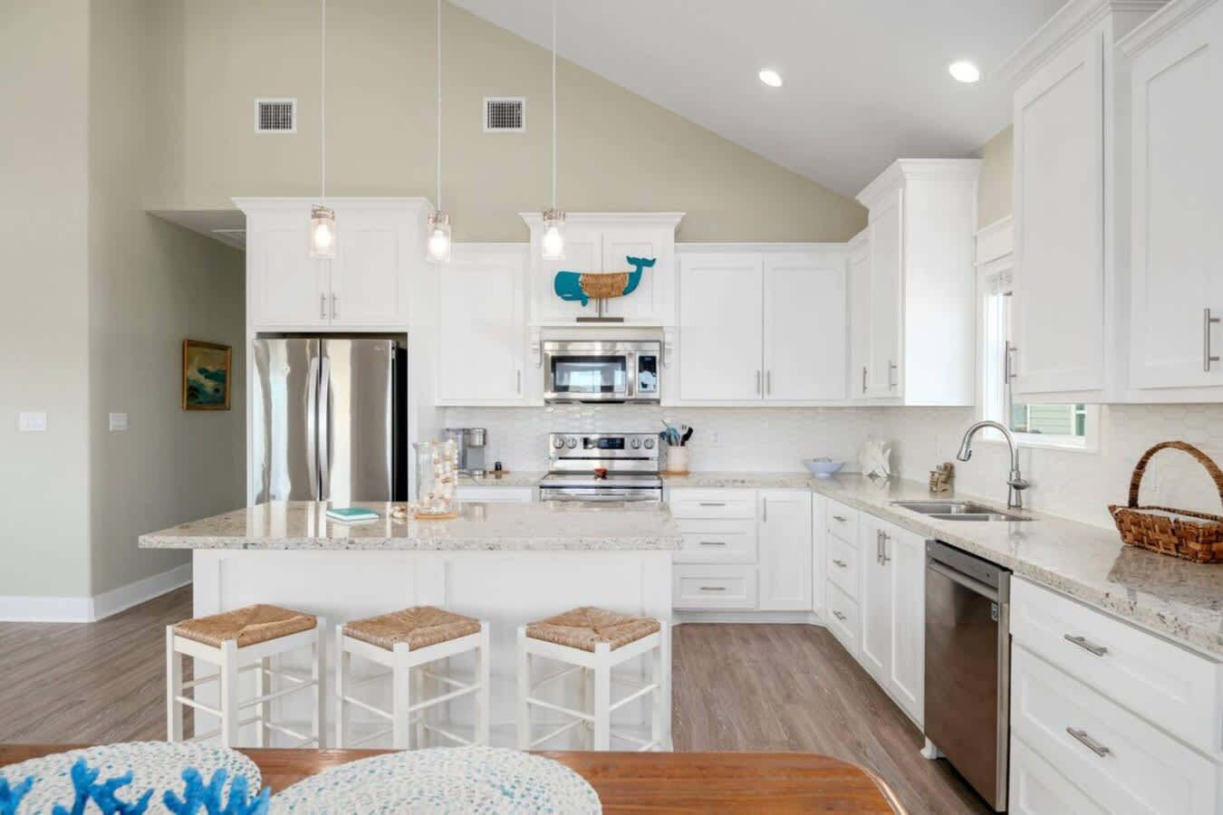 Una cocina moderna con armarios blancos, una isla central con taburetes, electrodomésticos de acero inoxidable e iluminación colgante.