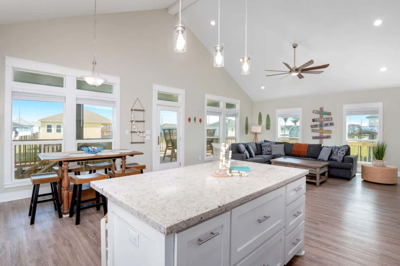 A spacious kitchen with a large island, a dining area with wooden chairs, a dark-colored sofa, and a ceiling fan.