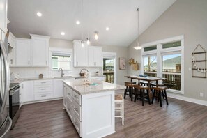 A modern kitchen with white cabinetry, a central island, and a dining area with wooden chairs. - 3 Bdr 3 Bth, King Bed, No Fees (Bolivar Peninsula)