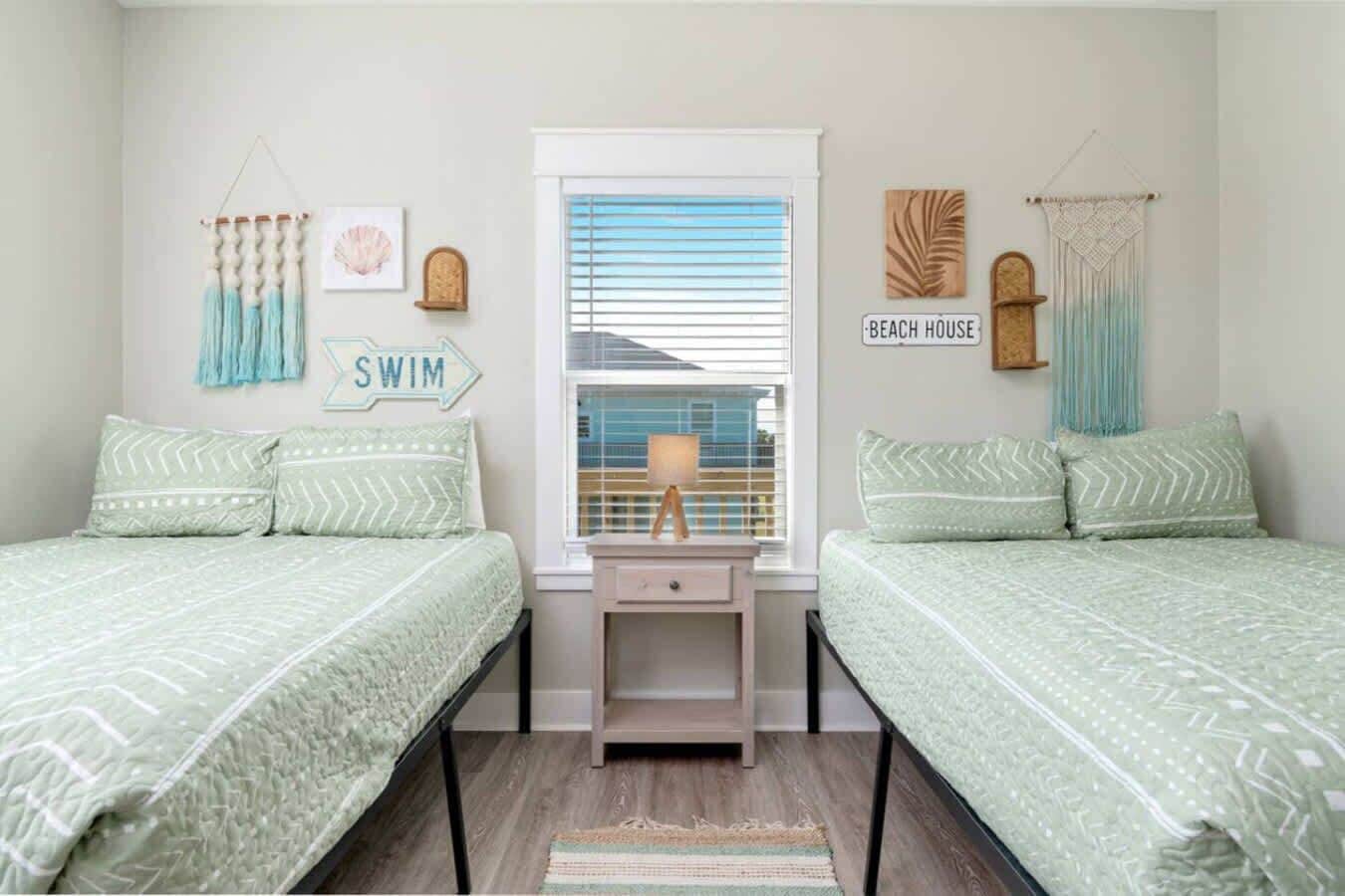 A bedroom with two beds, a window with blinds, a small table with a lamp, and wall decorations including a sign that says 'SWIM'.
