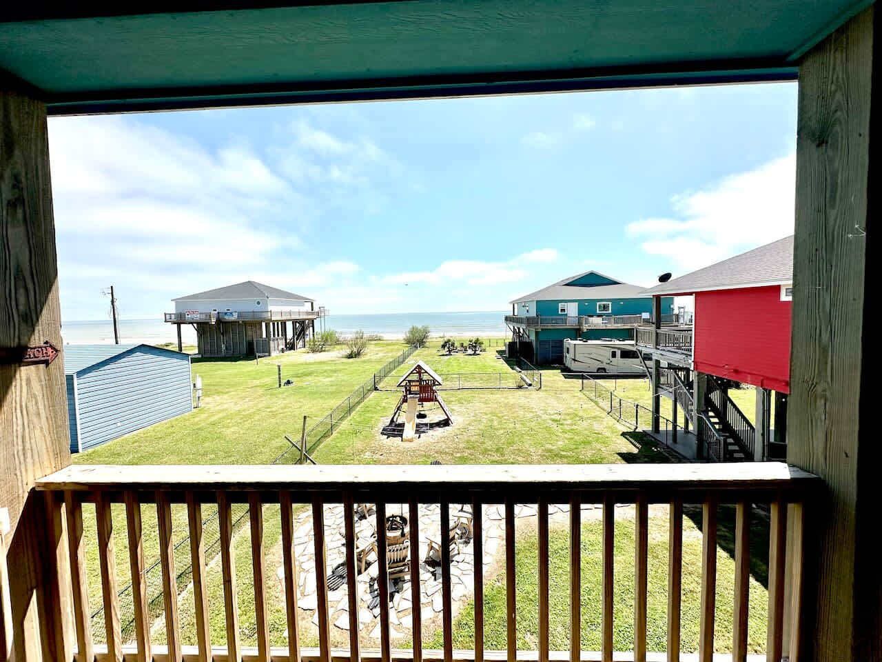 A view from a balcony overlooking a grassy area with a small play structure, a building with a red roof, and a blue building in the distance.