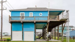A two-story building with a blue exterior, a balcony on the upper floor, and a garage on the lower floor. - Gulf Views, Steps to Beach, 2 King Beds, 3Bd 2Bth (Bolivar Peninsula)