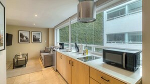 A modern kitchen with a microwave, sink, and cabinets, a window with a view of greenery, and a small seating area with a sofa. - Modern King Studio in Central Roma Norte (Ciudad de México)