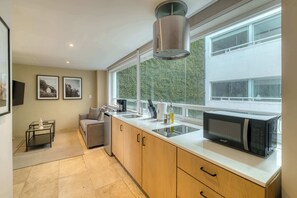 A modern kitchen with a microwave, sink, and cabinets, a window with a view of greenery, and a small seating area with a sofa. - Modern King Studio in Central Roma Norte (Ciudad de México)