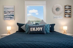 A bedroom with a bed, two bedside lamps, a window with blinds, a wall-mounted sign reading 'BEACH HOUSE', and a framed fish decoration. - 3 Bd 2 5 Bth, King Bed, Pet Friendly (Bolivar Peninsula)