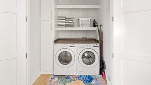 A laundry room with two front-loading washing machines, a shelf with folded towels, a laundry basket, and a mop. - Golf Cart, Elevator, Stylish, 12 guests - LaPerla (Bolivar Peninsula)