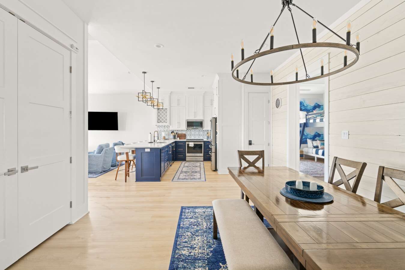 A modern kitchen with a central island, a dining area with a wooden table and chairs, and a hallway leading to a living room with a sofa and a TV.