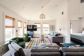 Ein modernes Wohnzimmer mit Fernseher, grünen und grauen Sofas, einem farbenfrohen StreifenTeppich und einer Decke mit einer besonderen Leuchte.