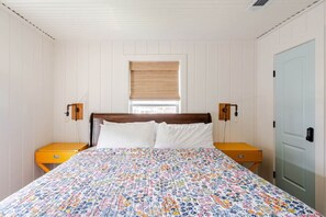 Ein Schlafzimmer mit einem Holz-Kopfende, einer Bettdecke mit Blumenmuster, zwei Nachttischen, einem Fenster mit Jalousien und einer Tür mit Schloss.