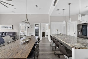 A modern kitchen with a wooden dining table, bar stools, and a built-in island with a refrigerator. - Bring the Whole Crew Sleeps 13 MerSea (Bolivar Peninsula)