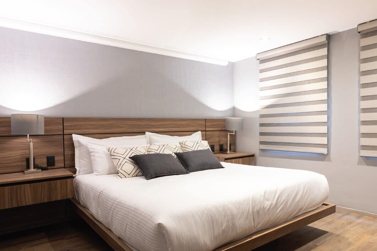 A modern bedroom with a wooden bed frame, white bedding, and a nightstand with a lamp.