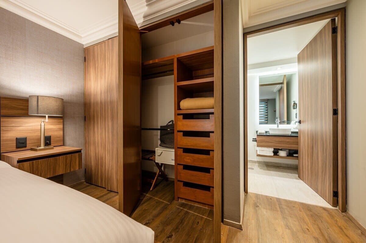 A bedroom with a wooden wardrobe, a bedside table with a lamp, and a view into a bathroom with a mirror and sink.