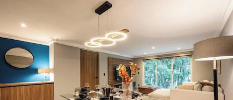 A modern dining area with a round table set for a meal, a large window with a view of greenery, and a contemporary light fixture above.