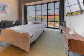 Ein modernes Hotelzimmer mit einem großen Bett, einem Schreibtisch mit Stühlen, einem Holznachttisch und Blick durch große Fenster.