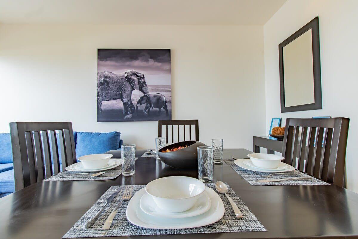 Une table à manger est dressée avec de l’assiette, des bols et des couverts blancs, et entourée de chaises à l’accoudoir recouvert d’un tissu sombre et agrémentées d’un coussin bleu.
