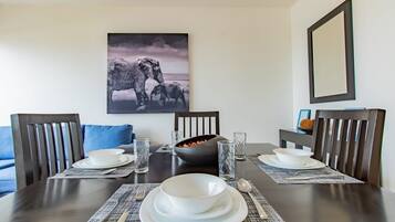 Une table à manger est dressée avec de l’assiette, des bols et des couverts blancs, et entourée de chaises à l’accoudoir recouvert d’un tissu sombre et agrémentées d’un coussin bleu.