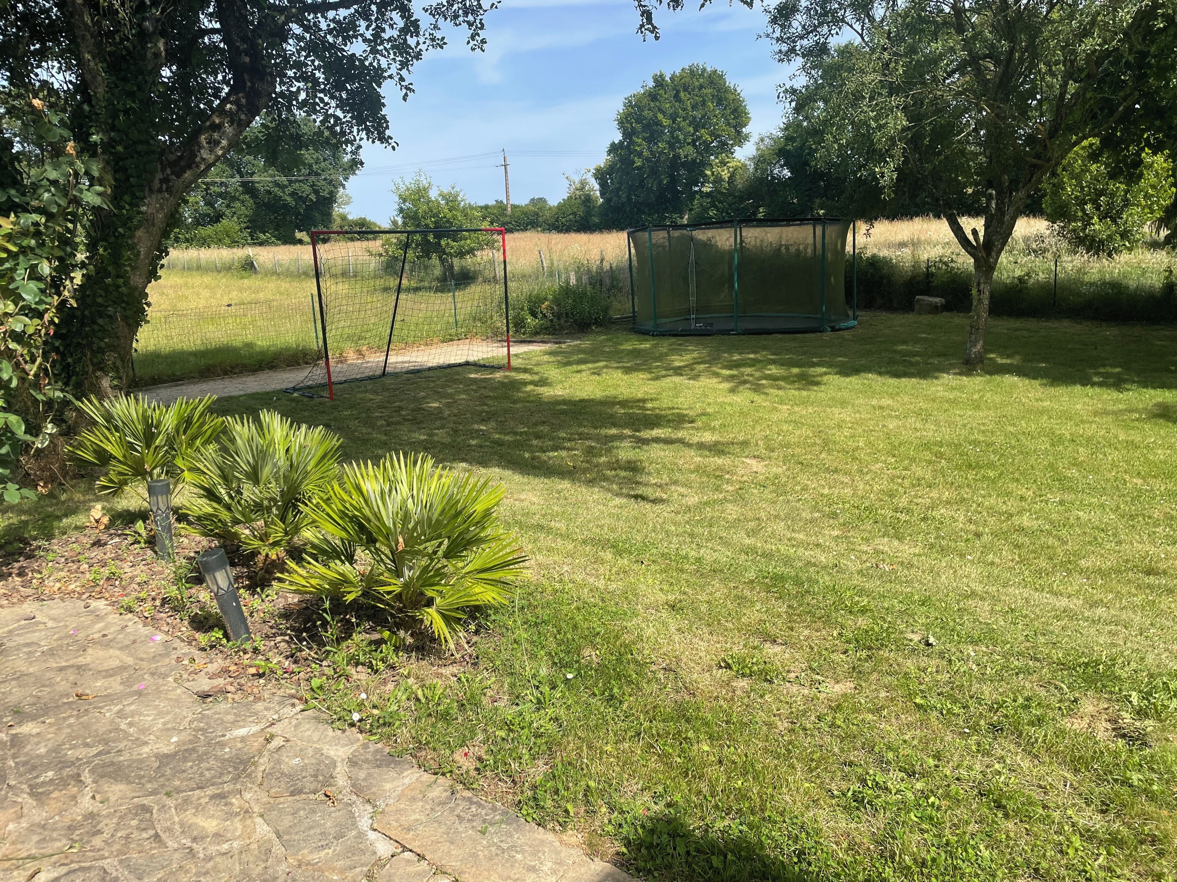 Un espace extérieur verdoyant avec une aire de jeux clôturée, des arbres et un sentier en pierre.