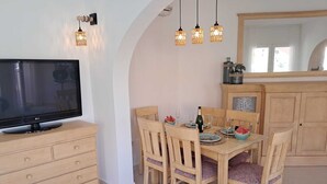 A dining area with a TV on a cabinet, wooden chairs, a table set for two, and hanging pendant lights. - Villa in Cumbre del Sol—bookable for stays of 11 days or more (El Poble Nou de Benitatxell/Benitachell)